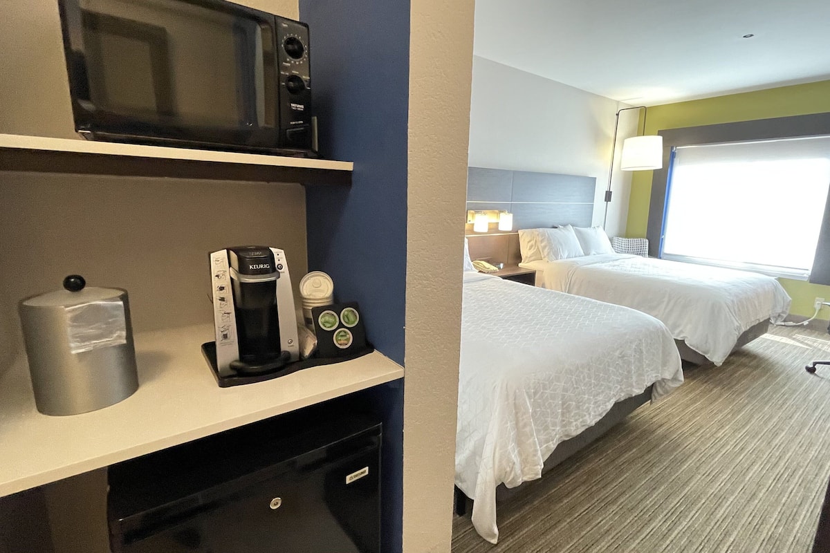 A hotel room with a kitchenette, two beds, a window with a view, and a desk with a coffee maker.