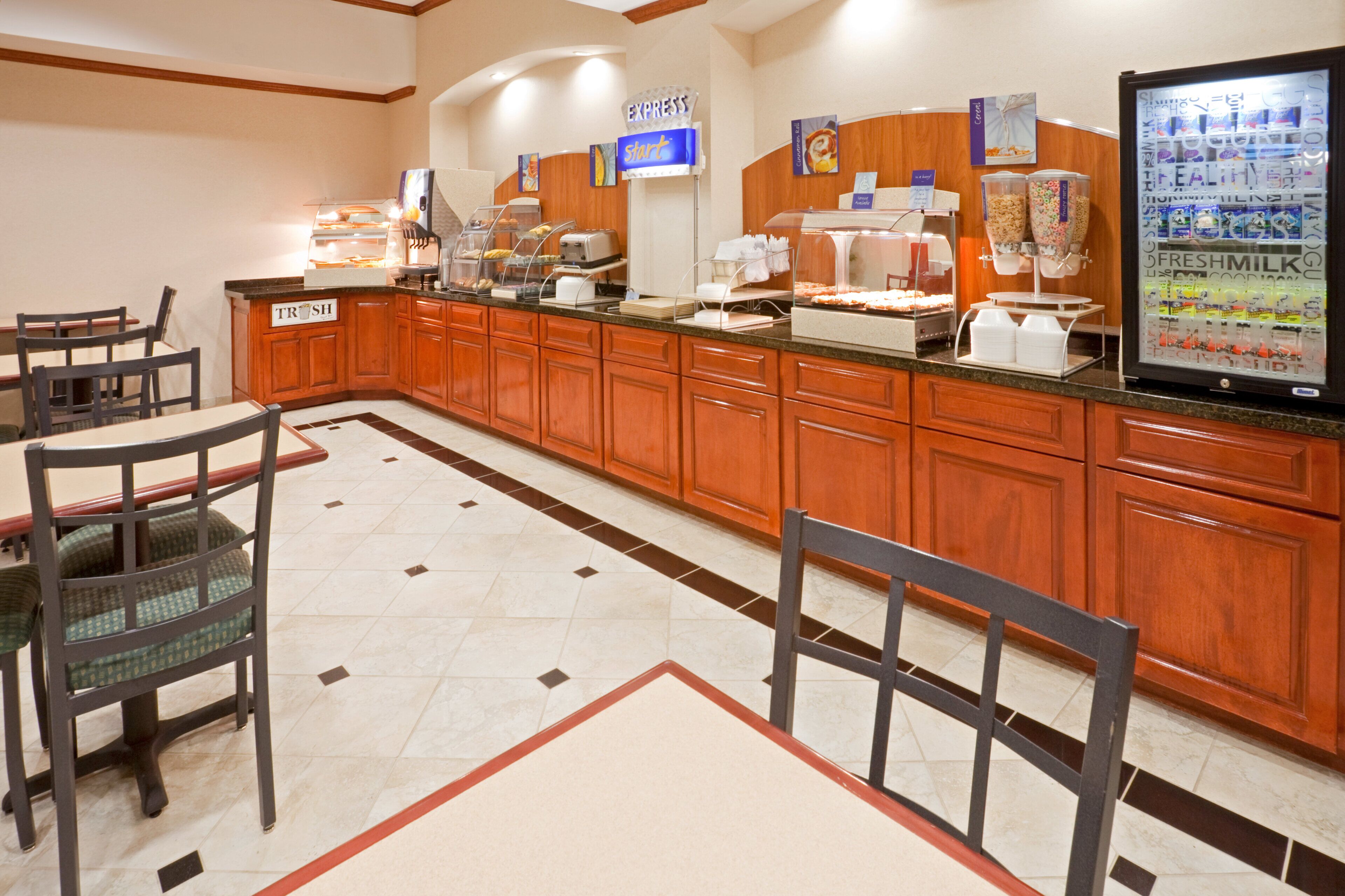 A hotel breakfast area with a buffet counter, seating, and a refrigerator displaying food items.