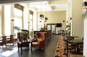 A spacious hotel lobby with a long reception desk, multiple seating areas, and a large TV screen.