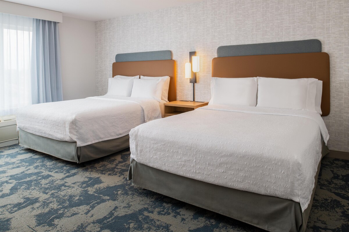Habitación de hotel con dos camas, una mesita de noche con lámpara y una alfombra estampada.