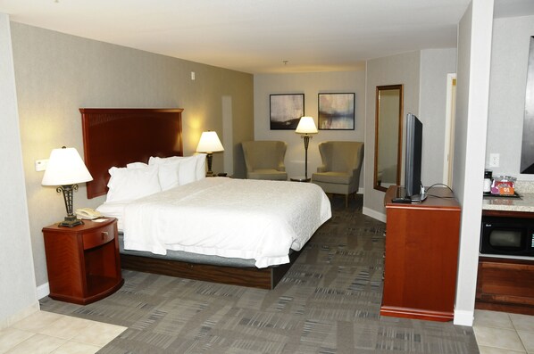 A hotel room with a large bed, two chairs, a desk with a TV, and a mirror.