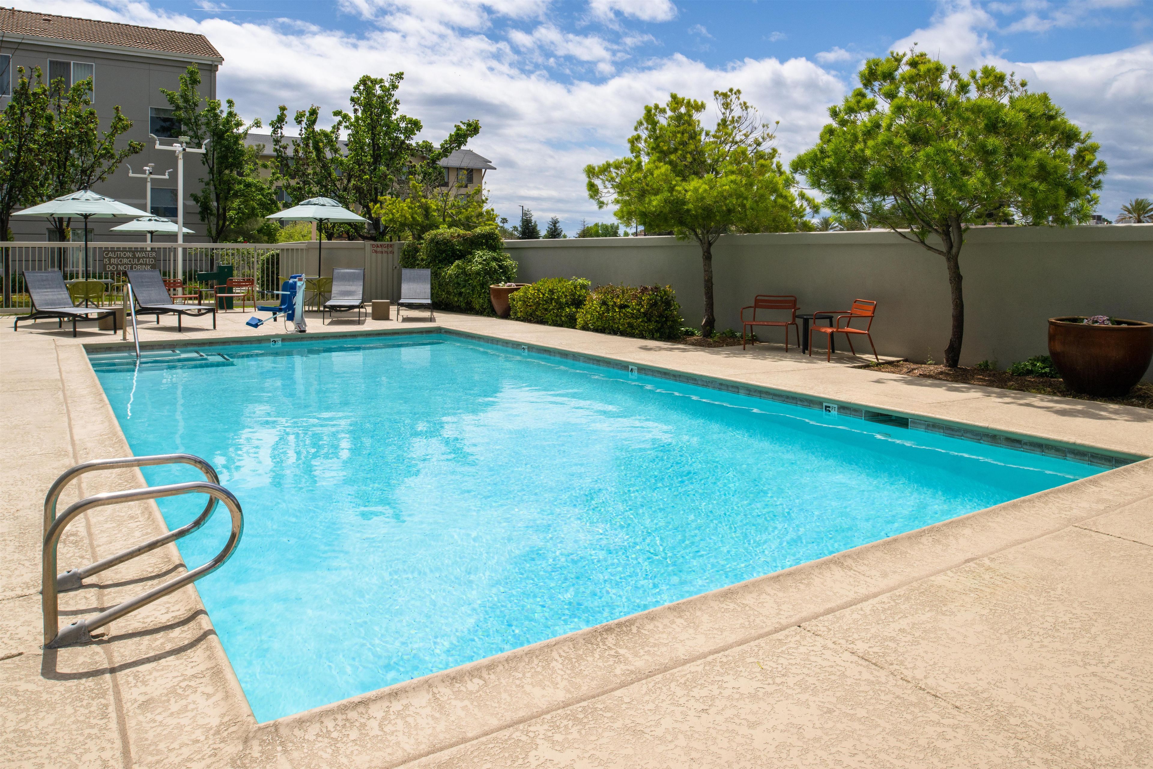 A rectangular swimming pool with a metal ladder, surrounded by lounge chairs and umbrellas, with a building and trees in the background.