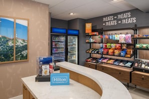 Una hall d'albergo con un banco reception, un minibar, un frigorifero ed scaffali pieni di snack.