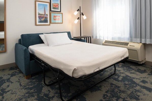 A single bed with a mattress, a blue upholstered headboard, a bedside table, a floor lamp, and a window with curtains.