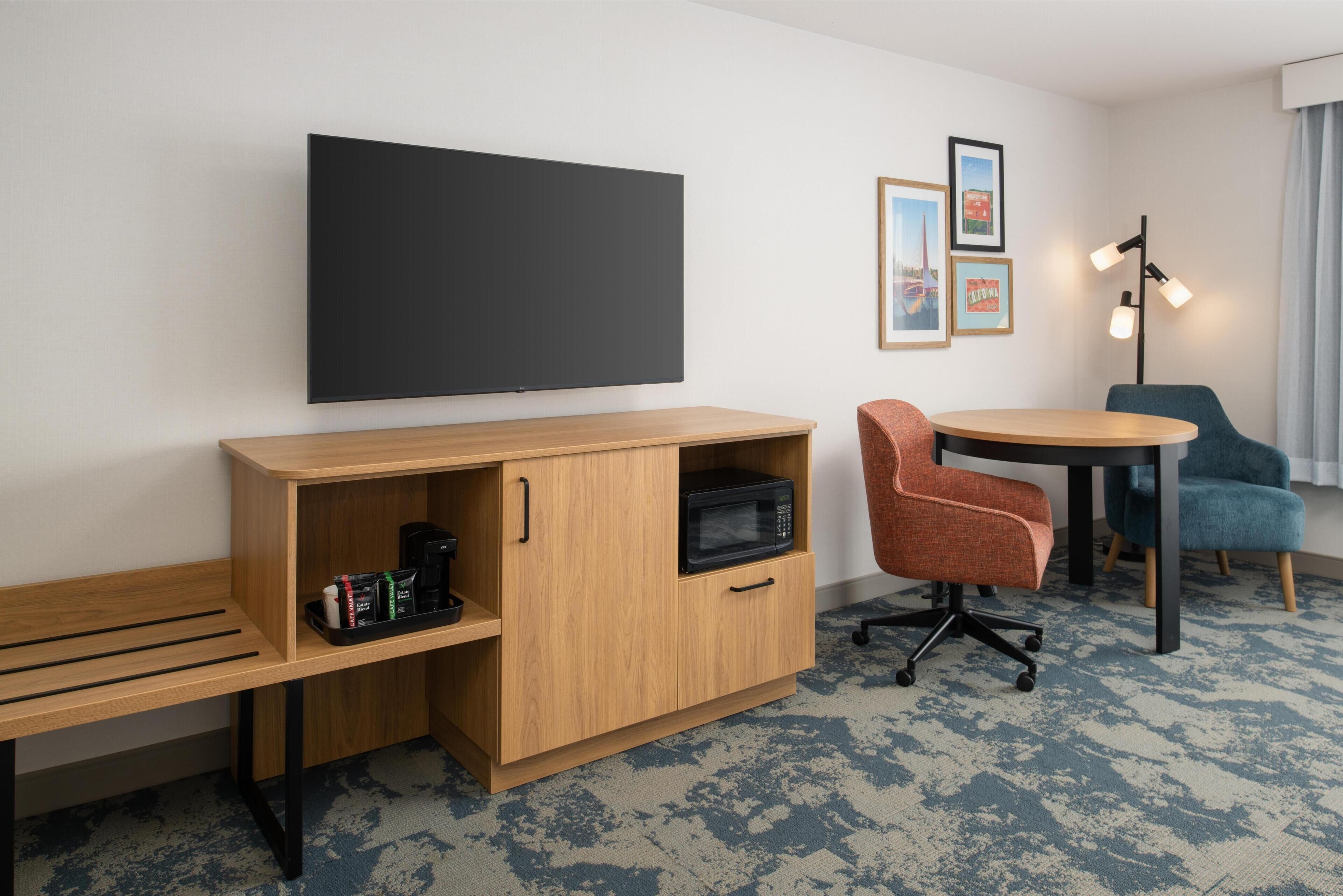 A hotel room with a flat-screen TV, wooden desk, office chair, small round table, and a floor lamp.