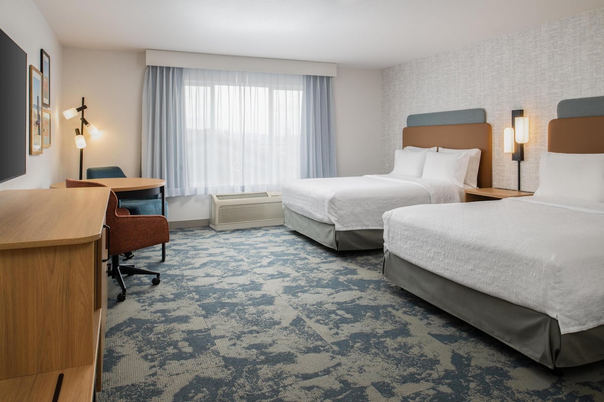 A hotel room with two beds, a desk, a chair, a window with curtains, and a wall-mounted lamp.
