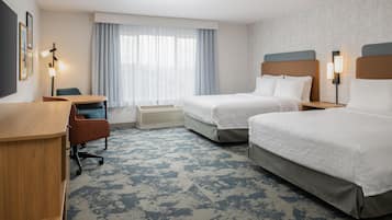 Quarto de hotel com duas camas, uma escrivaninha, uma cadeira, uma janela com cortinas e uma luminária na parede.