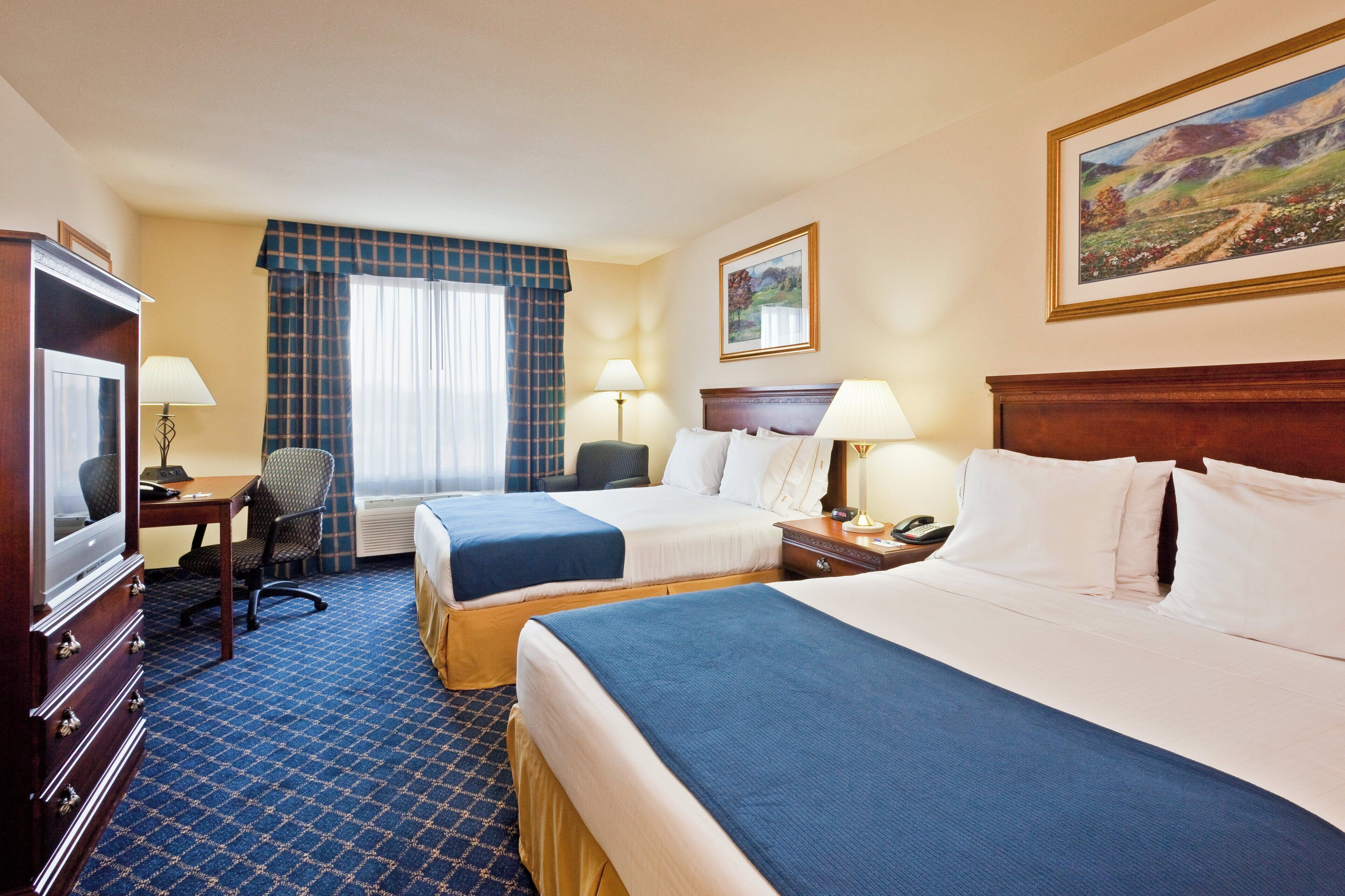 A hotel room with two beds, a desk with a television, a chair, a window with curtains, and a painting on the wall.
