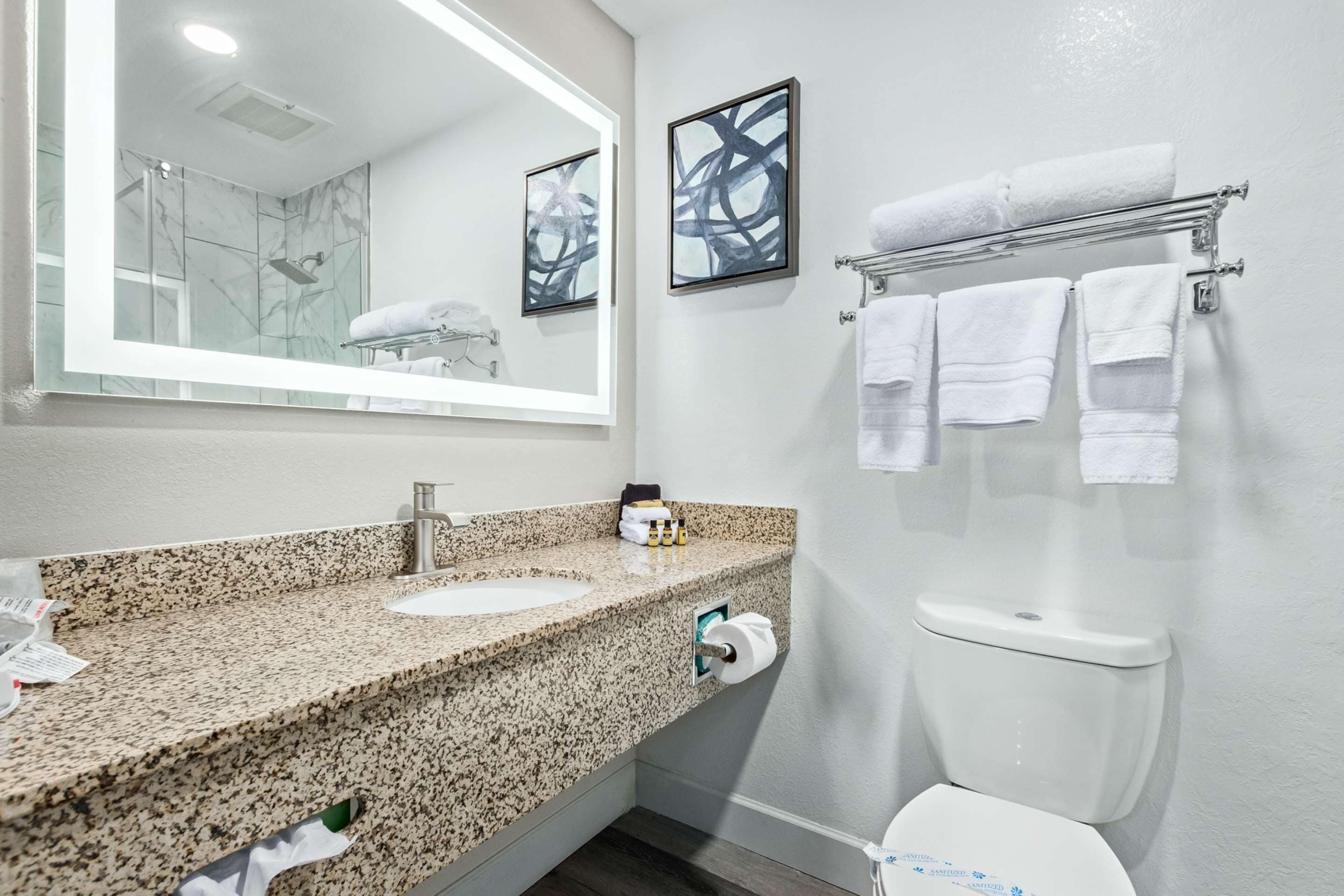 A bathroom with a granite countertop sink, a mirror, a toilet, and a towel rack with multiple towels.