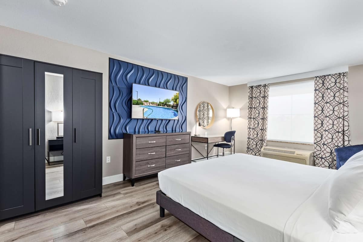 A modern hotel room with a large bed, a desk, a TV mounted on a wavy wall, and a view of a swimming pool.