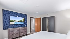 Quarto de hotel com uma cama grande, cômoda com gavetas, guarda-roupa e televisão na parede exibindo vista para a piscina.