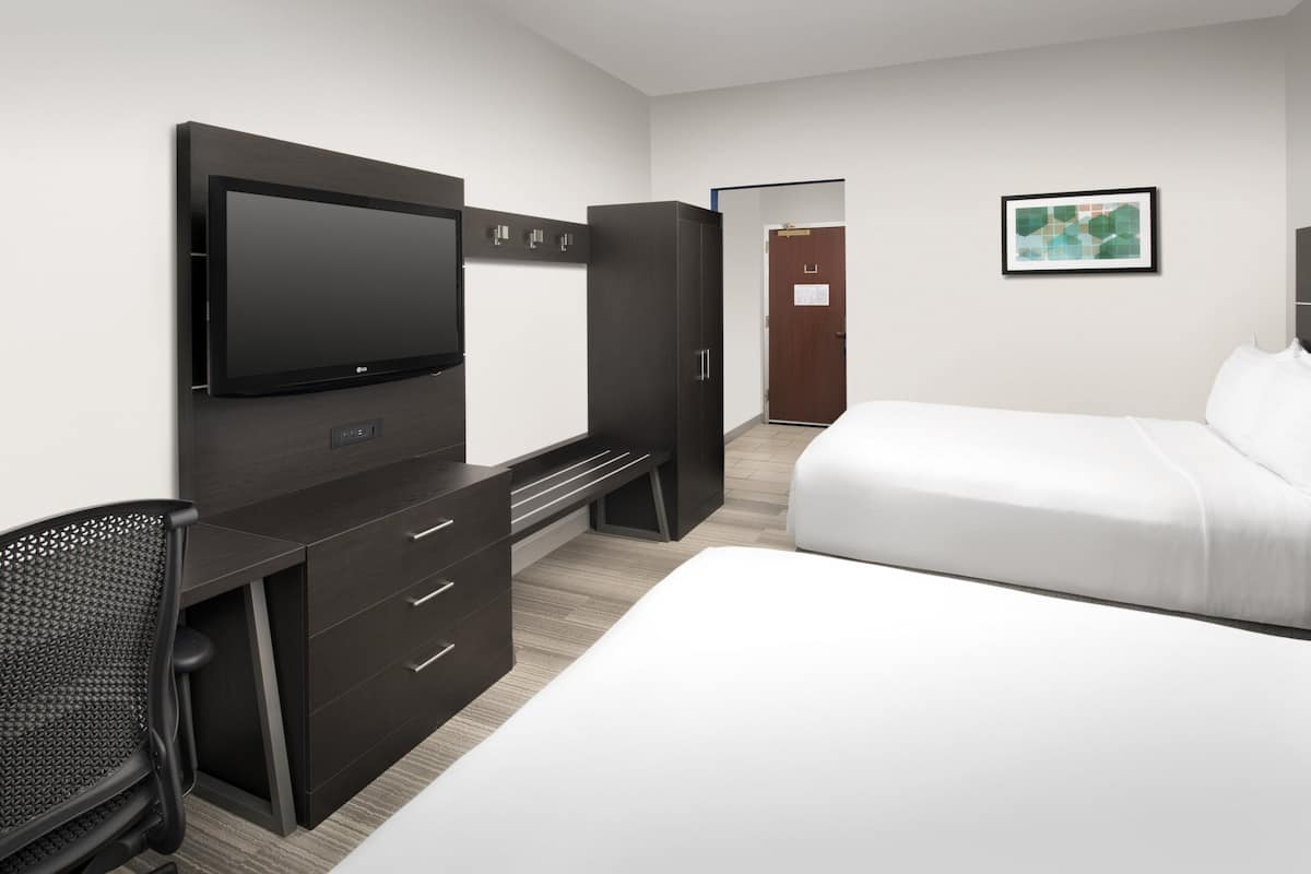 A modern hotel room with a flat-screen TV, a desk with drawers, a black office chair, a large bed with white linens, and a framed abstract art piece on the wall.