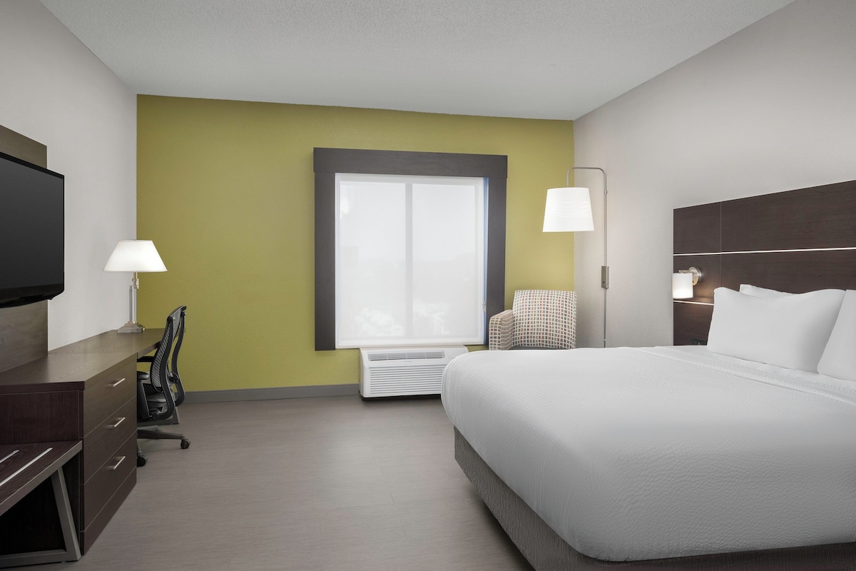 A hotel room with a bed, a desk, a chair, a TV, a window, and a lamp.