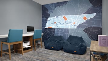 Una habitación con un escritorio, dos sillas, una impresora, un mural con un mapa y una estantería con libros y juegos.