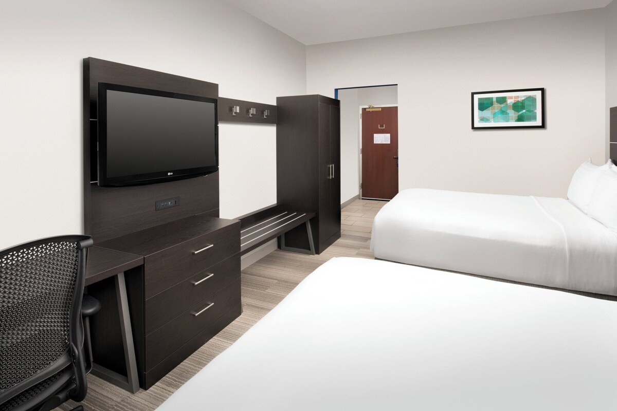 Une chambre d’hôtel moderne équipée d’un téléviseur à écran plat, d’un bureau avec des tiroirs, d’un fauteuil de bureau noir, d’un grand lit avec du linge de lit blanc et d’une œuvre d’art abstraite encadrée accrochée au mur.