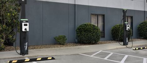 Un estacionamiento con dos estaciones de carga para vehículos eléctricos, un edificio de fachada gris y un pequeño arbusto.