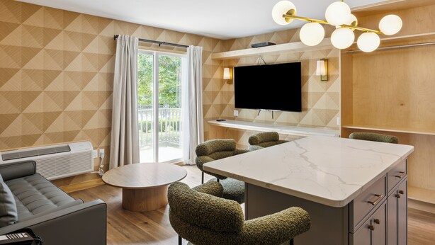 Un salon moderne comprenant un canapé, une table basse ronde et un espace cuisine doté d’un grand îlot, d’un téléviseur à écran plat et d’un luminaire original.