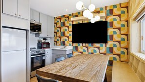 A modern kitchen with white cabinets, a built-in oven, and a microwave. There's a wooden dining table with chairs, a TV mounted on the wall, and a patterned wallpaper.