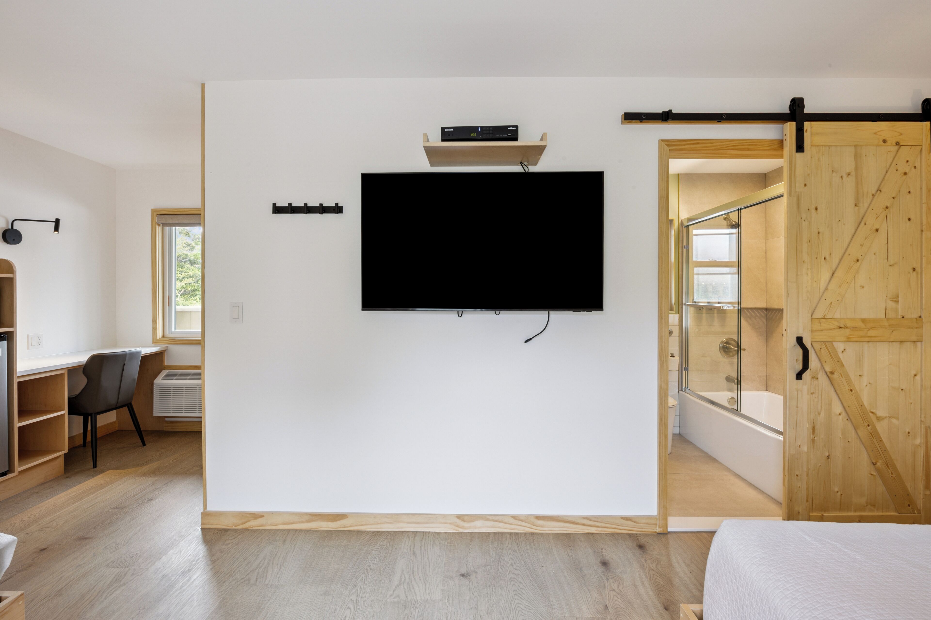 Ein modernes Zimmer mit montiertem Fernseher, Holzboden, einem Schreibtisch mit Stuhl und einer Schiebetür vom Typtennbretter, die ins Badezimmer führt.