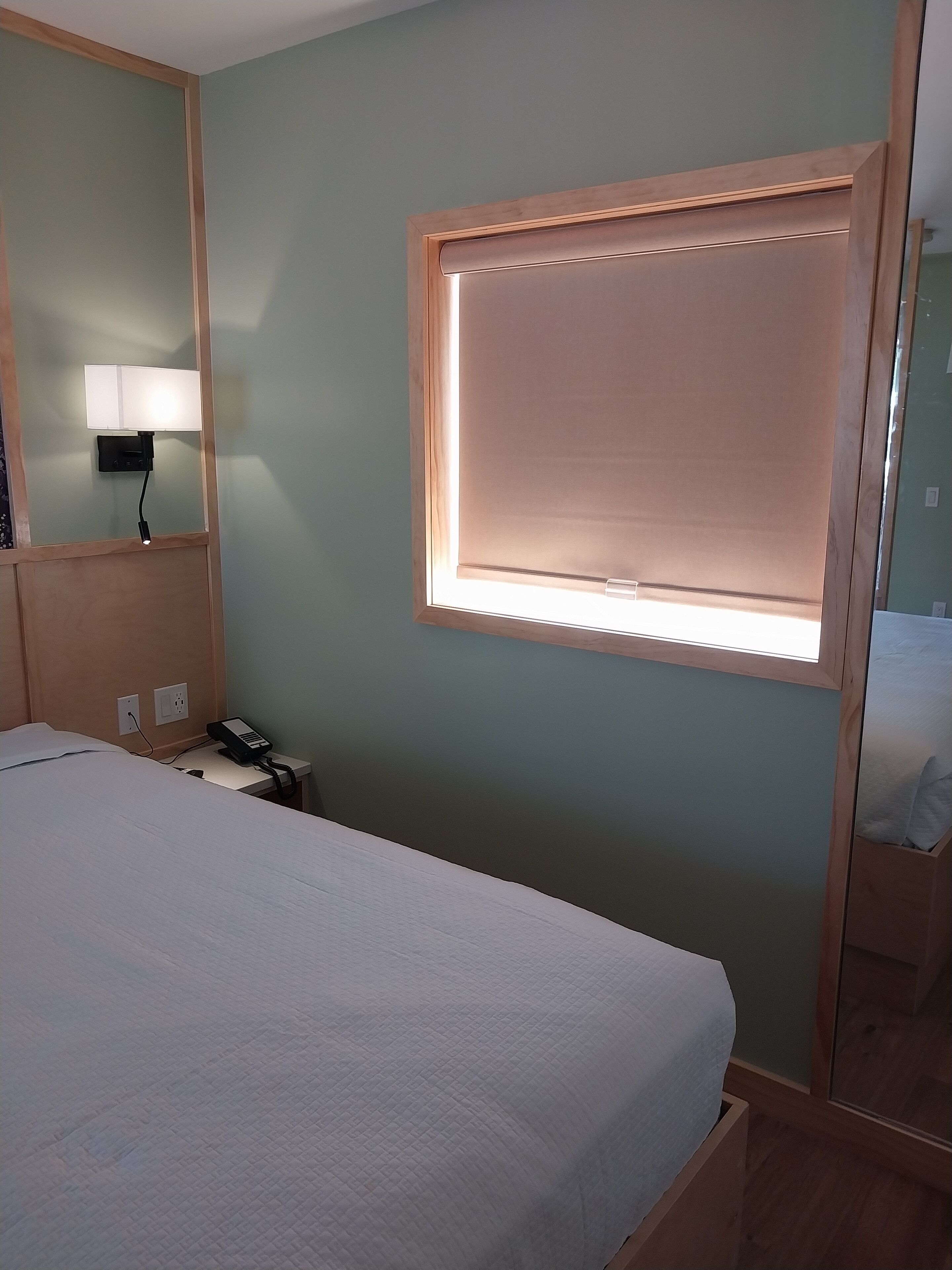 Een hotelkamer met een bed, nachtkastje, lamp en een raam met zonwering.