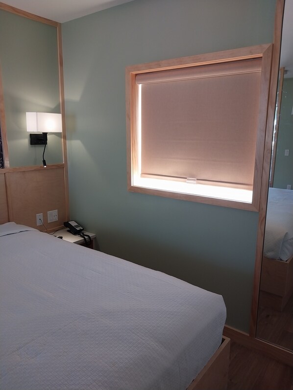 A hotel room with a bed, bedside table, lamp, and a window with blinds.