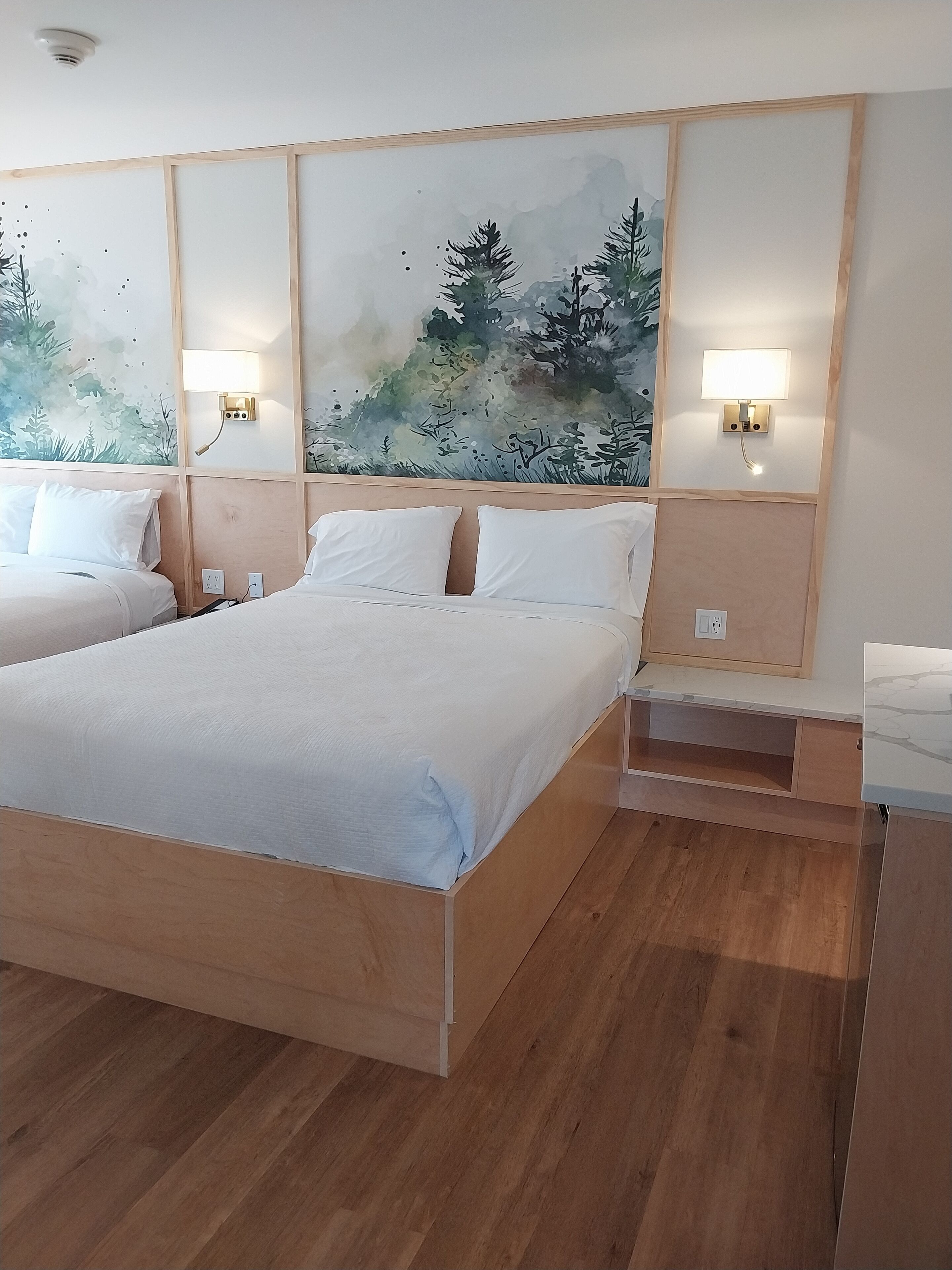 Habitación de hotel con una cama grande, una cama más pequeña, cabecero de madera, dos lámparas de pared y un cuadro con una escena de bosque.