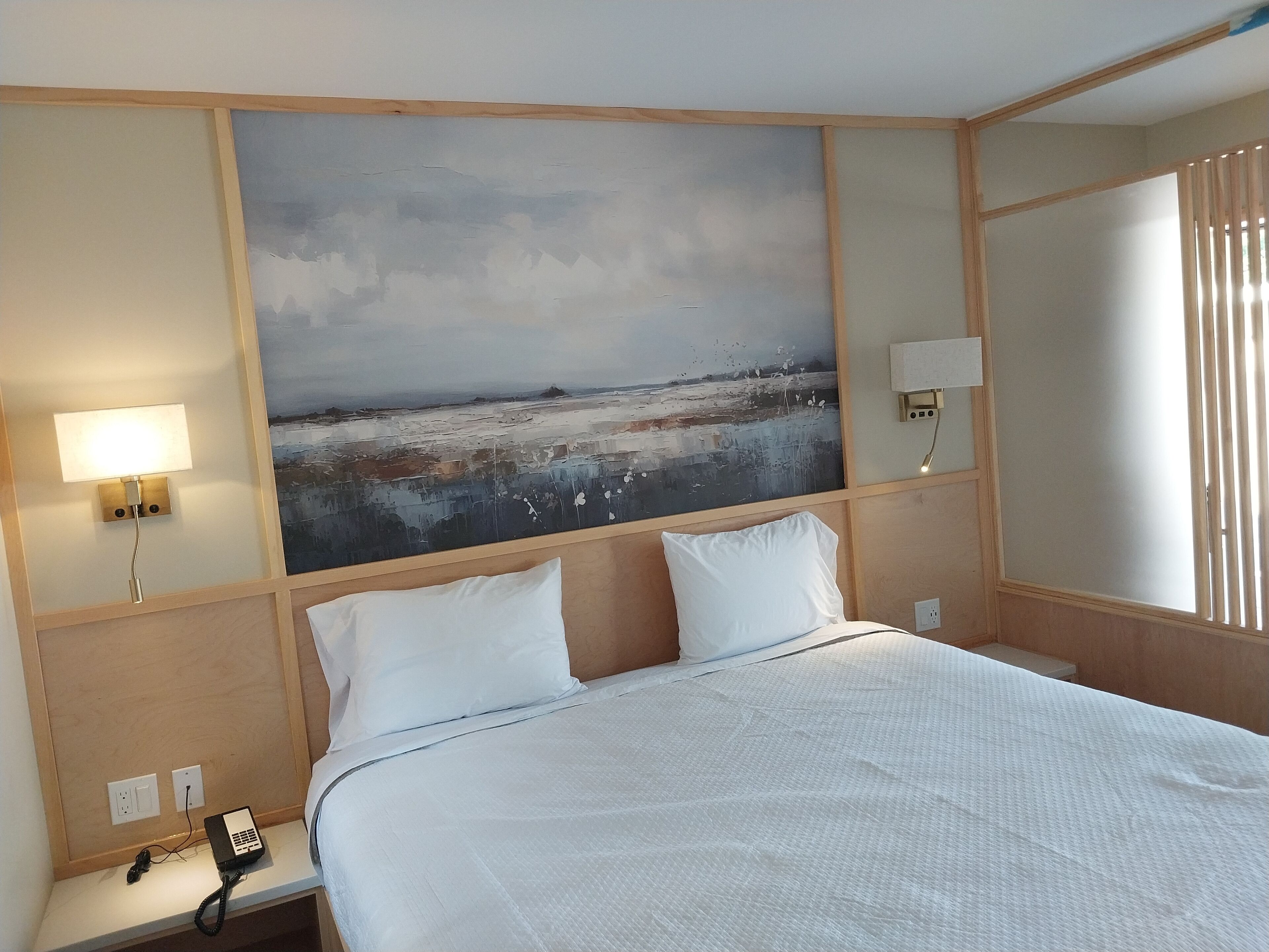 A hotel room with a bed, a painting on the wall, a nightstand with a lamp, and a phone.