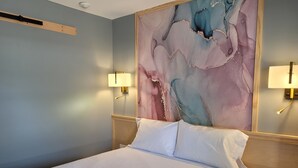 A hotel room with a bed, two wall-mounted lamps, a large abstract painting, and a wooden headboard.