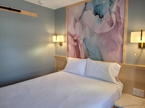 A hotel room with a bed, two wall-mounted lamps, a large abstract painting, and a wooden headboard.