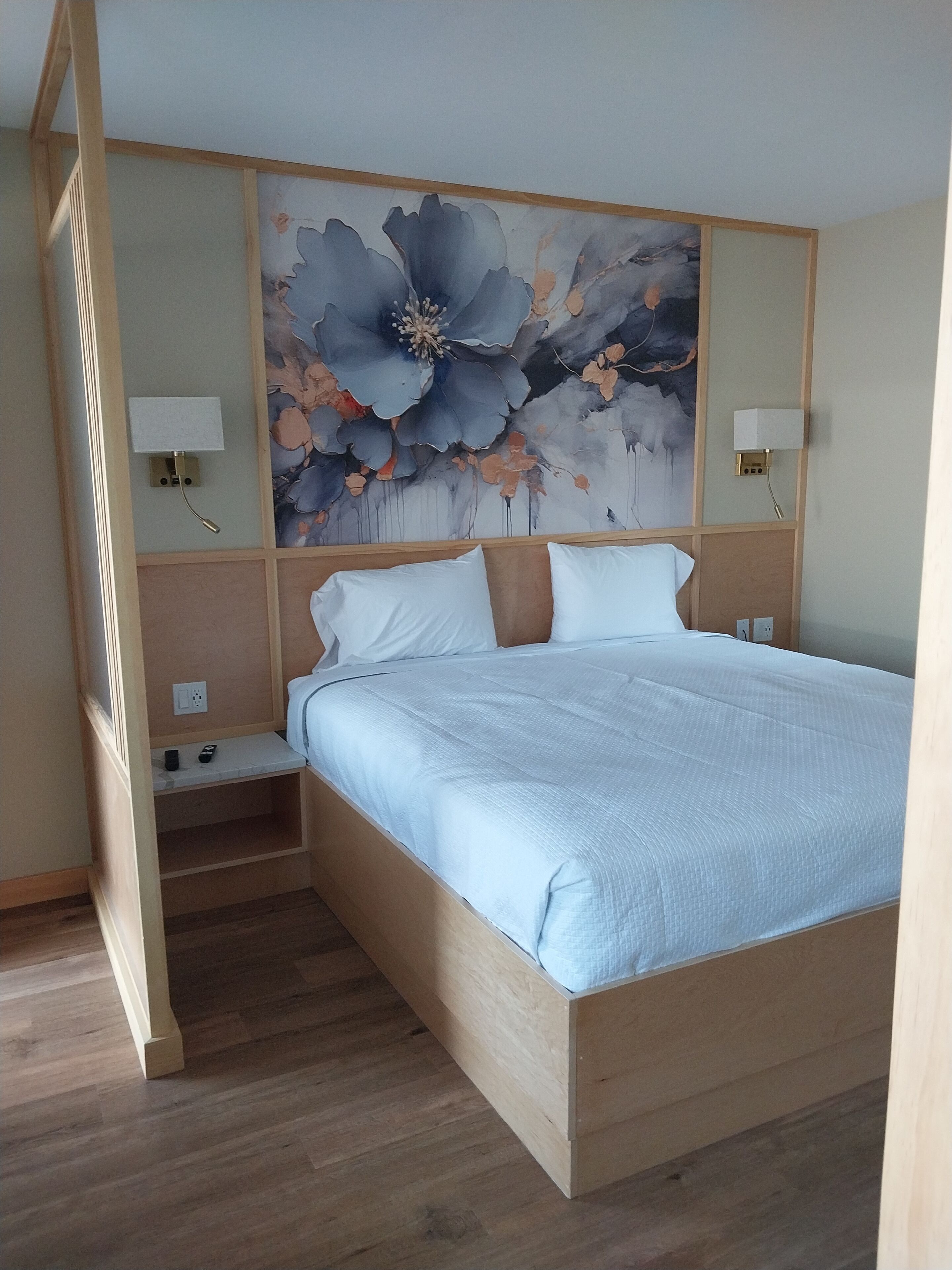 Une chambre à coucher avec un lit en bois, du linge de lit blanc, une grande œuvre d’art florale accrochée au mur et des lampes de chevet.