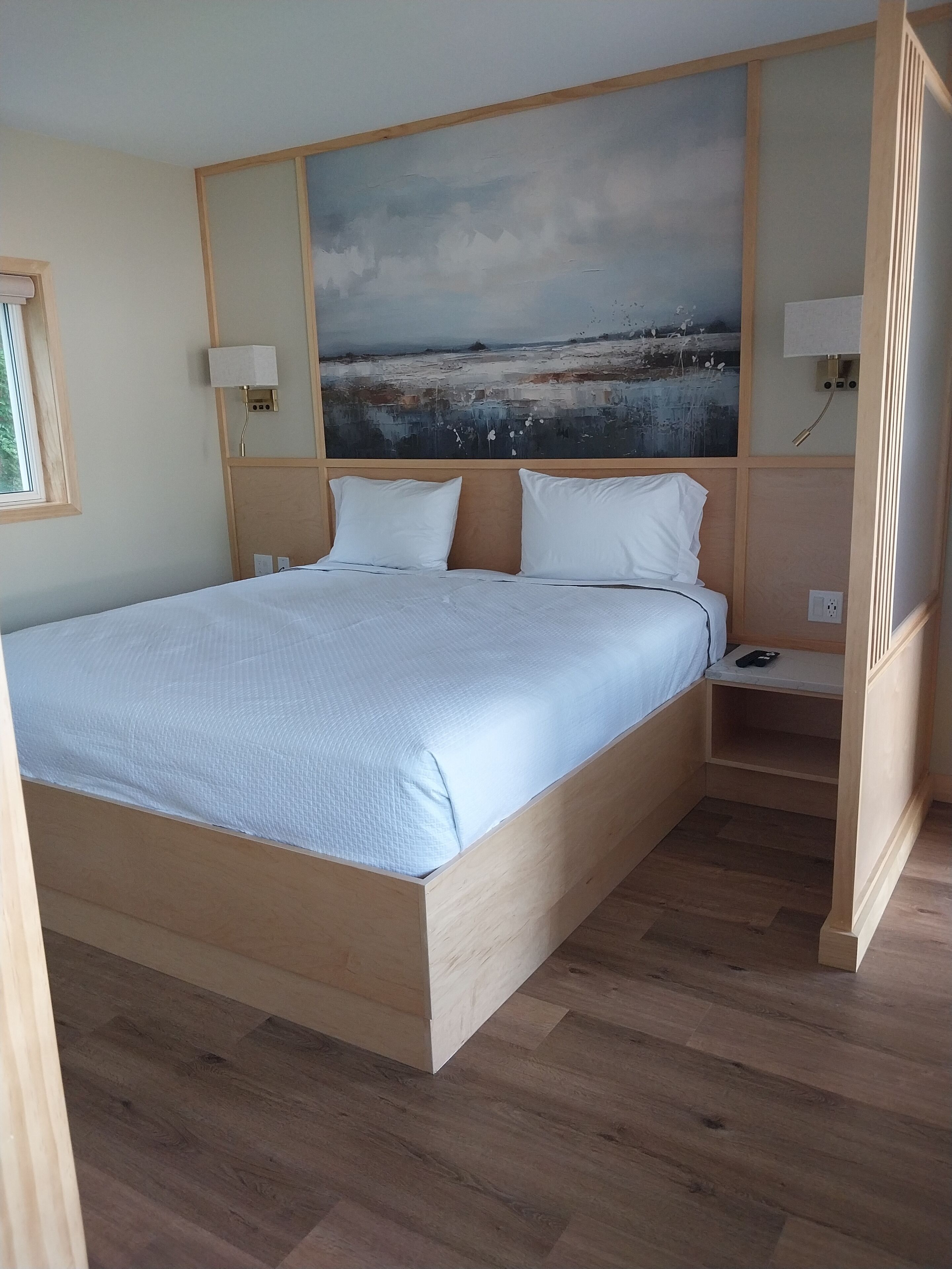 Un dormitorio con una cama de madera, una mesita de noche, una lámpara de pared y un cuadro de paisaje grande.