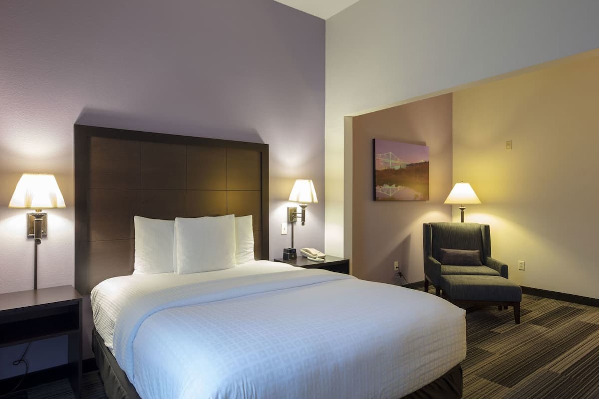 Ein Hotelzimmer mit einem Bett, zwei Nachttischlampen, einem Sessel, einem Gemälde an der Wand und einem Telefon auf dem Nachttisch.