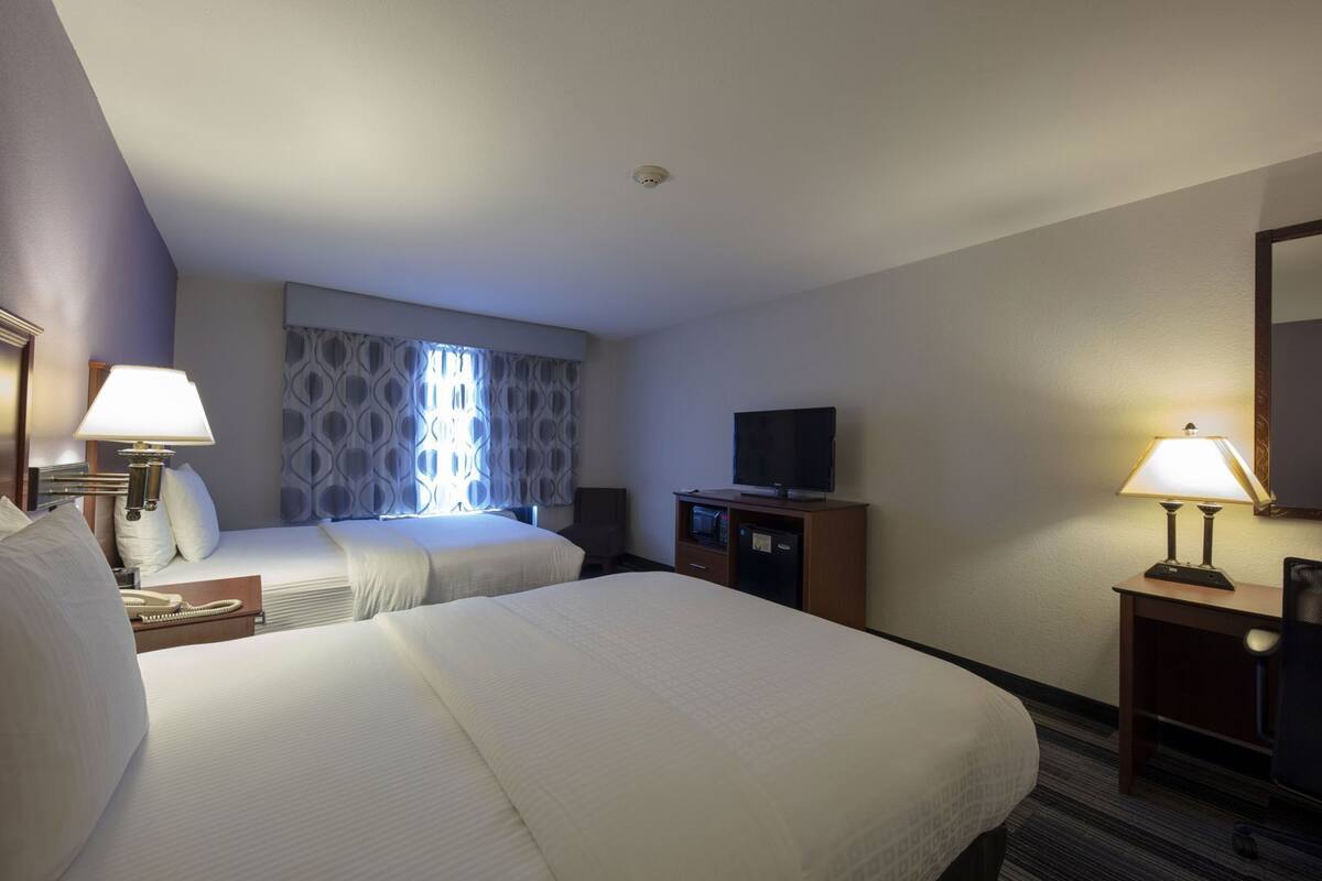 Ein Hotelzimmer mit zwei Betten, einem Fernseher, einem Schreibtisch mit Lampe und einem Spiegel.