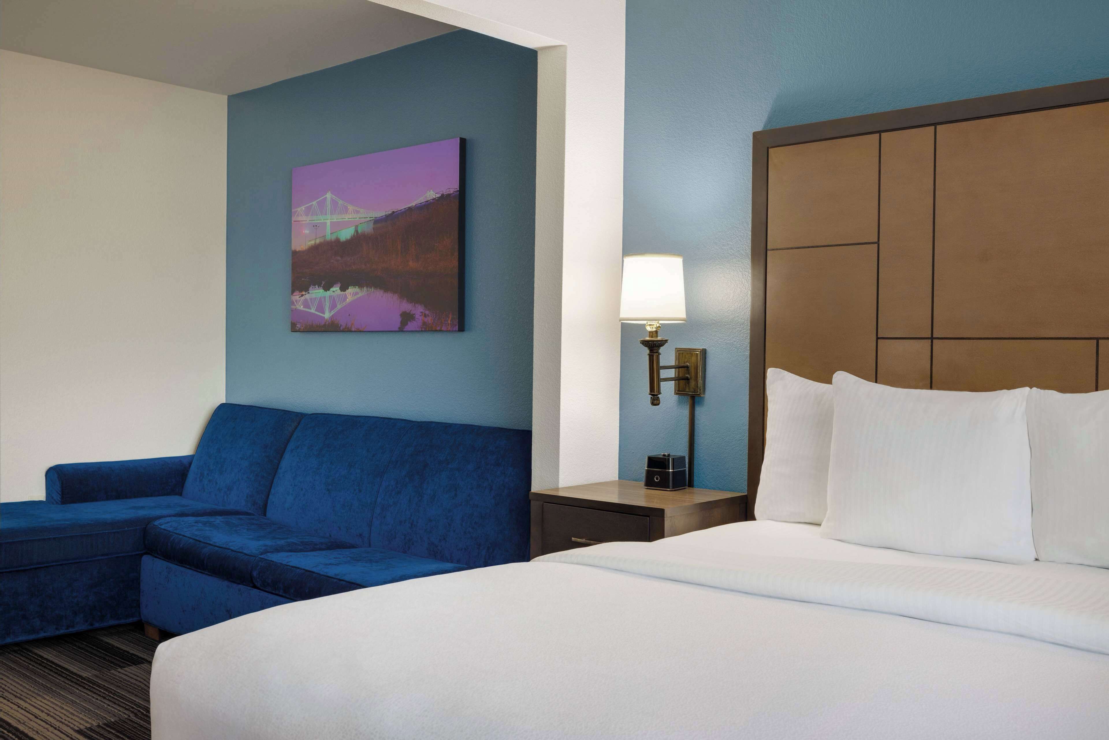 A hotel room with a large bed, a blue sofa, a nightstand with a lamp, and a wall-mounted painting.