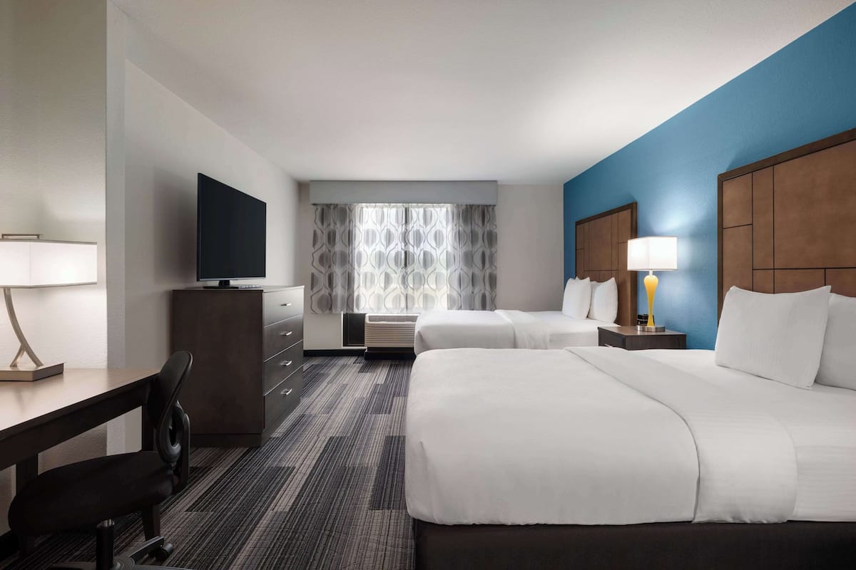 Ein Hotelzimmer mit Bett, Schreibtisch mit Stuhl, Fernseher, Fenster mit gemusterten Vorhängen und Nachttisch mit Lampe.