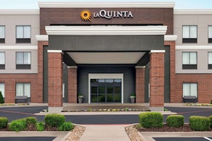 Fachada de hotel La Quinta con diseño moderno, que muestra un logotipo destacado, un acceso cubierto y un frente ajardinado con bancos y plantas en macetas.