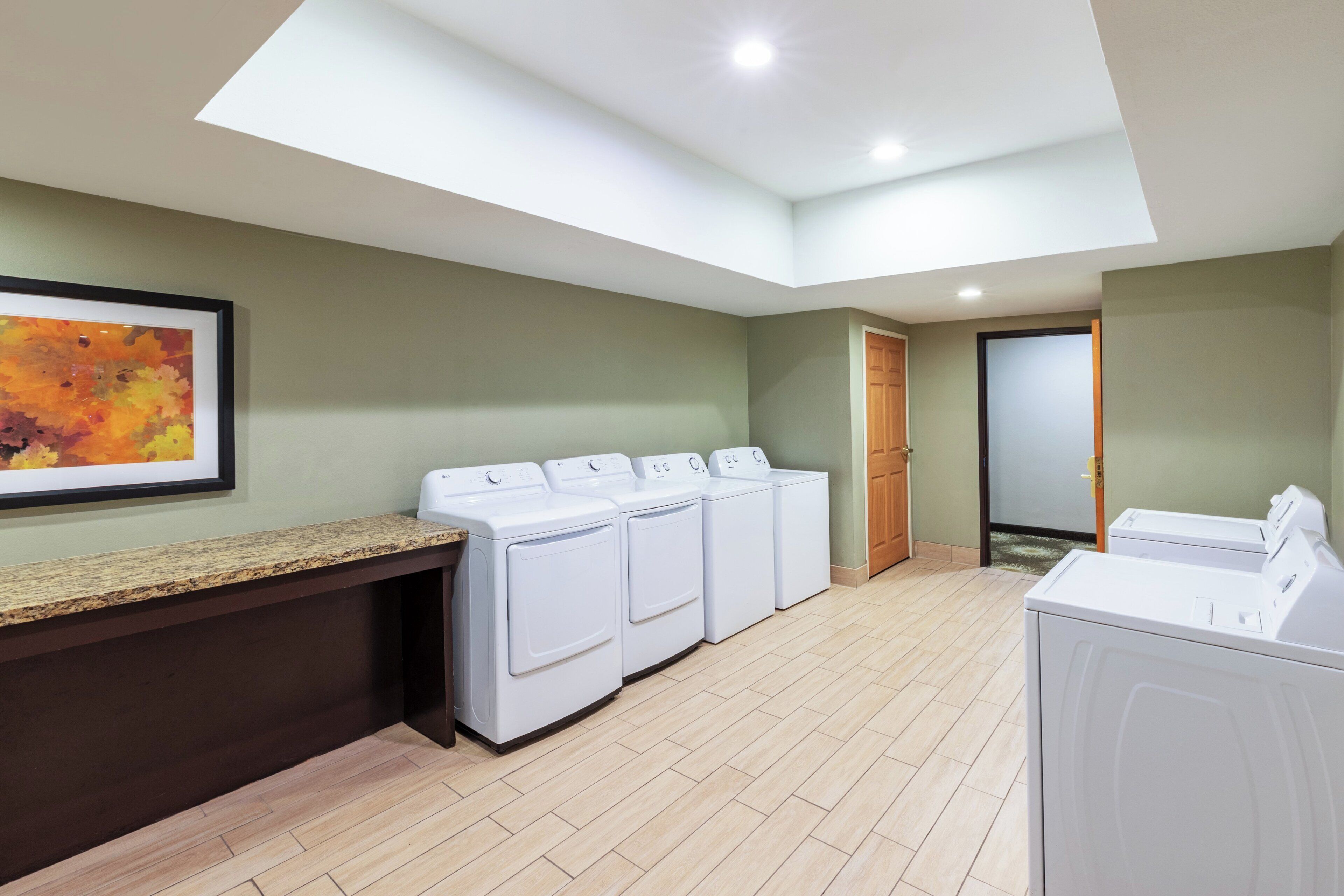 A laundry room with a countertop, four washing machines, a wooden door, and a framed artwork on the wall.