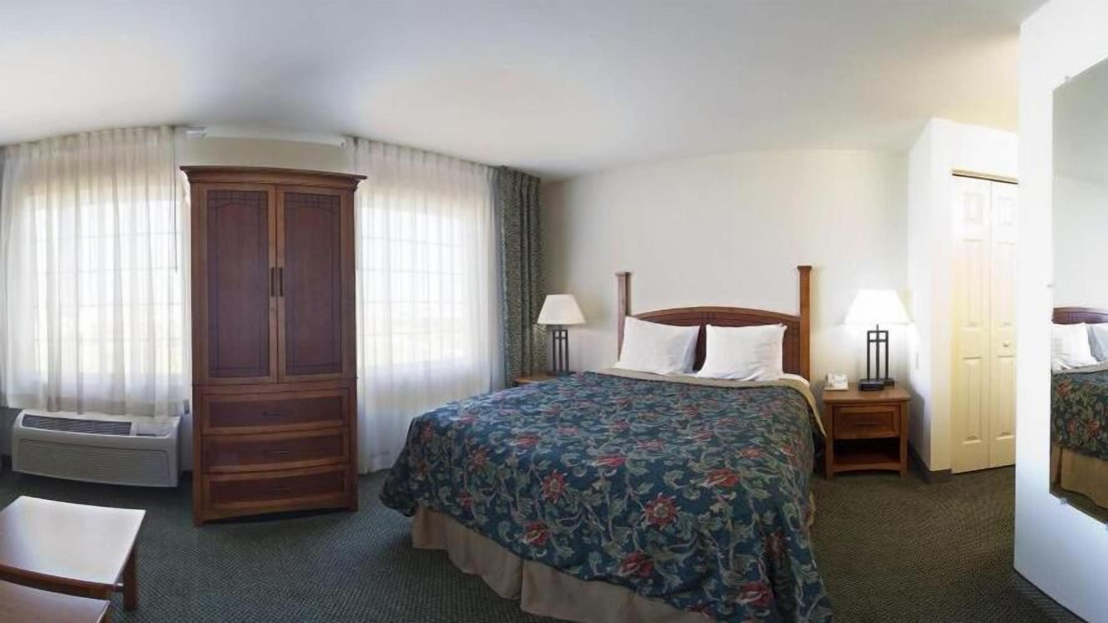 Una habitación de hotel con una cama grande, un armario, un escritorio con lámpara y un espejo.