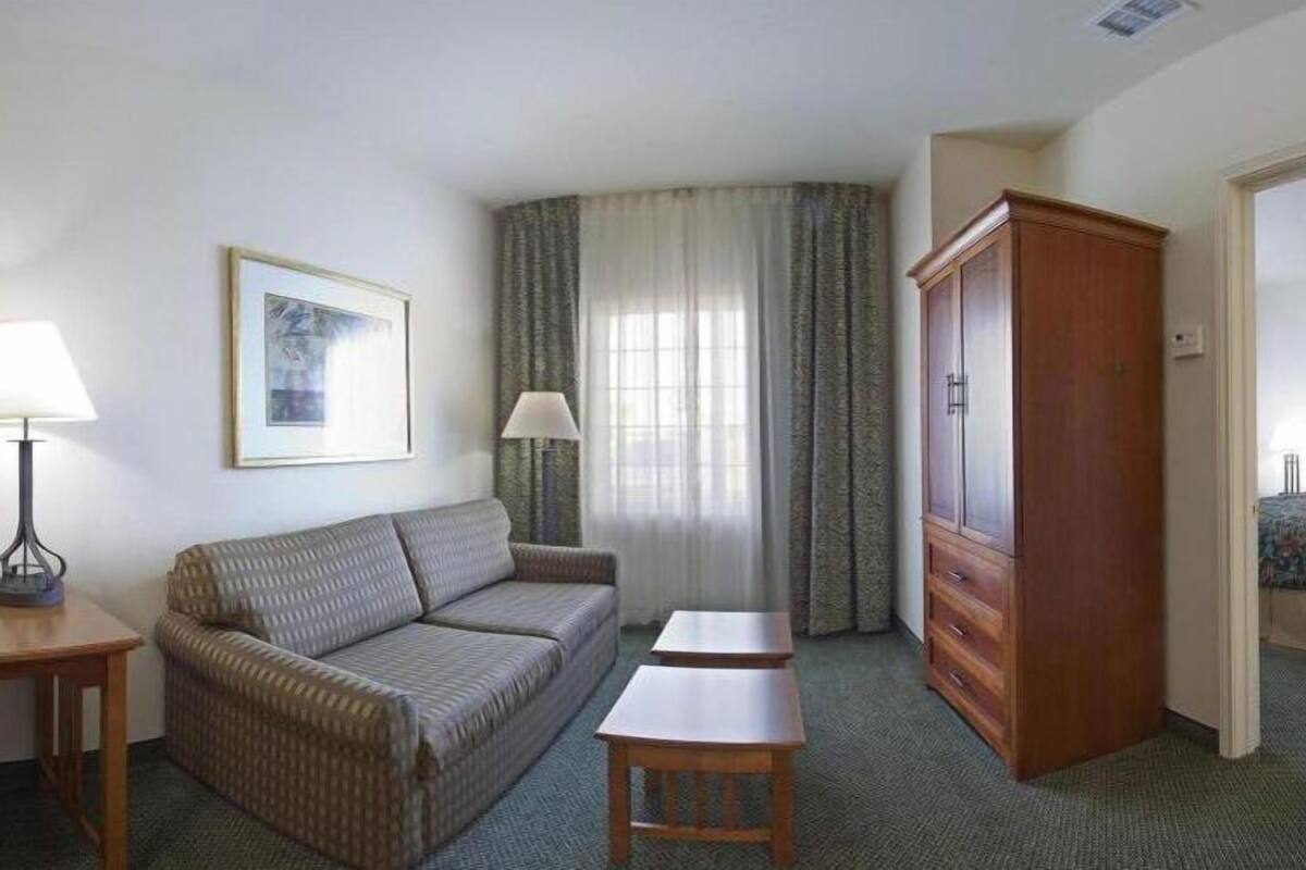 Una habitación de hotel con sofá, dos mesitas, un armario, cama y ventana con cortinas.