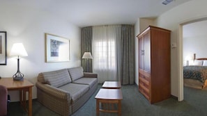 Ein Hotelzimmer mit einer Couch, zwei kleinen Tischen, einem Schrank, einem Bett und einem Fenster mit Vorhängen.