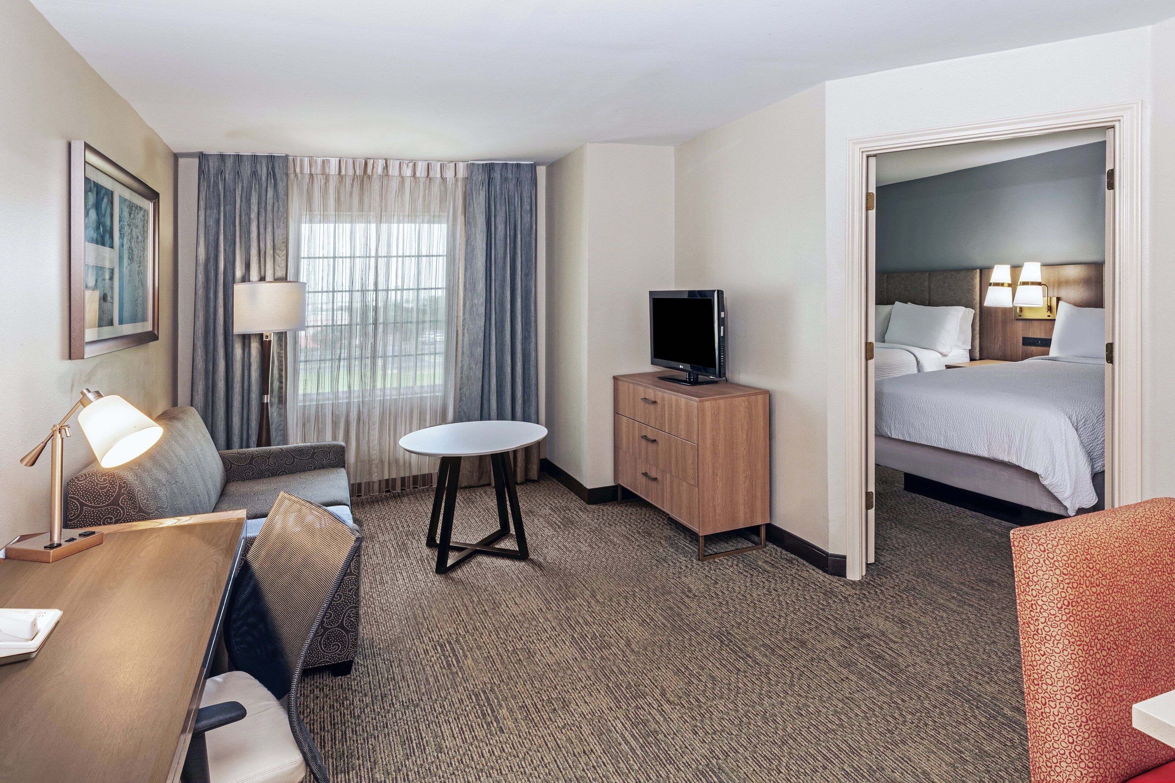 Habitación de hotel con cama, escritorio, silla, televisión y ventana con cortinas.