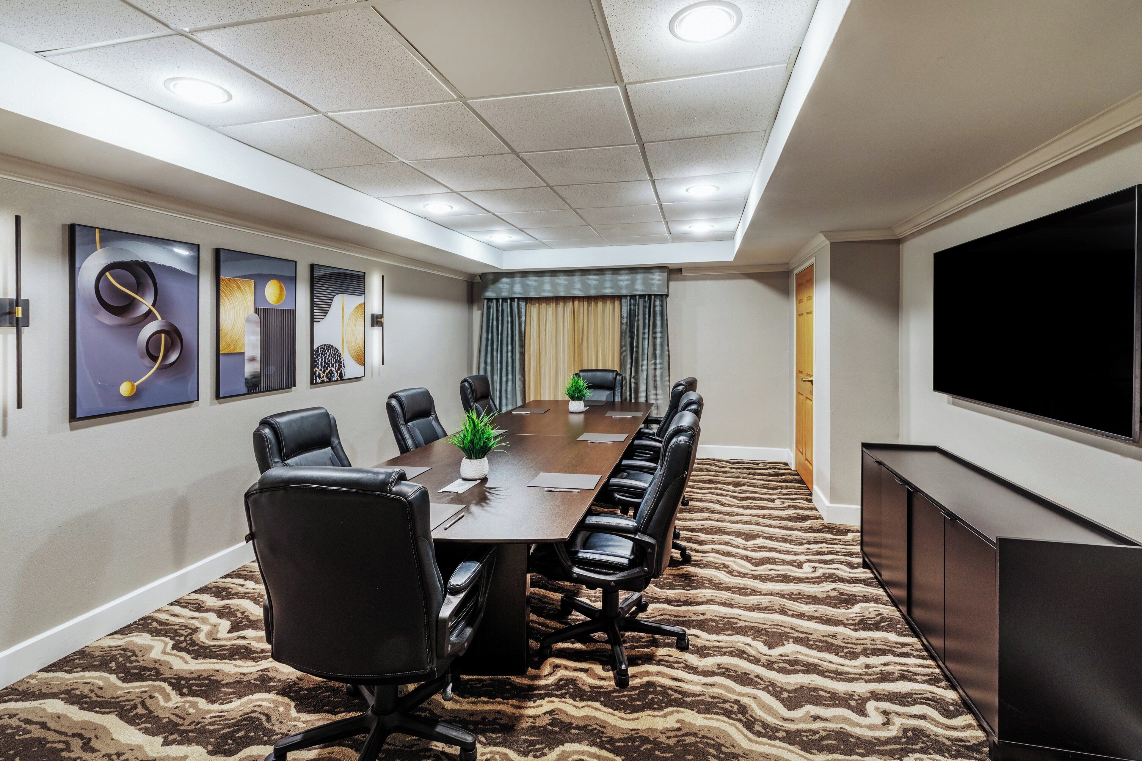 A conference room with a long table, black leather chairs, a projector screen, and a television on the wall.