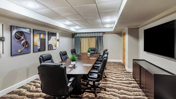 A conference room with a long table, black leather chairs, a projector screen, and a television on the wall.