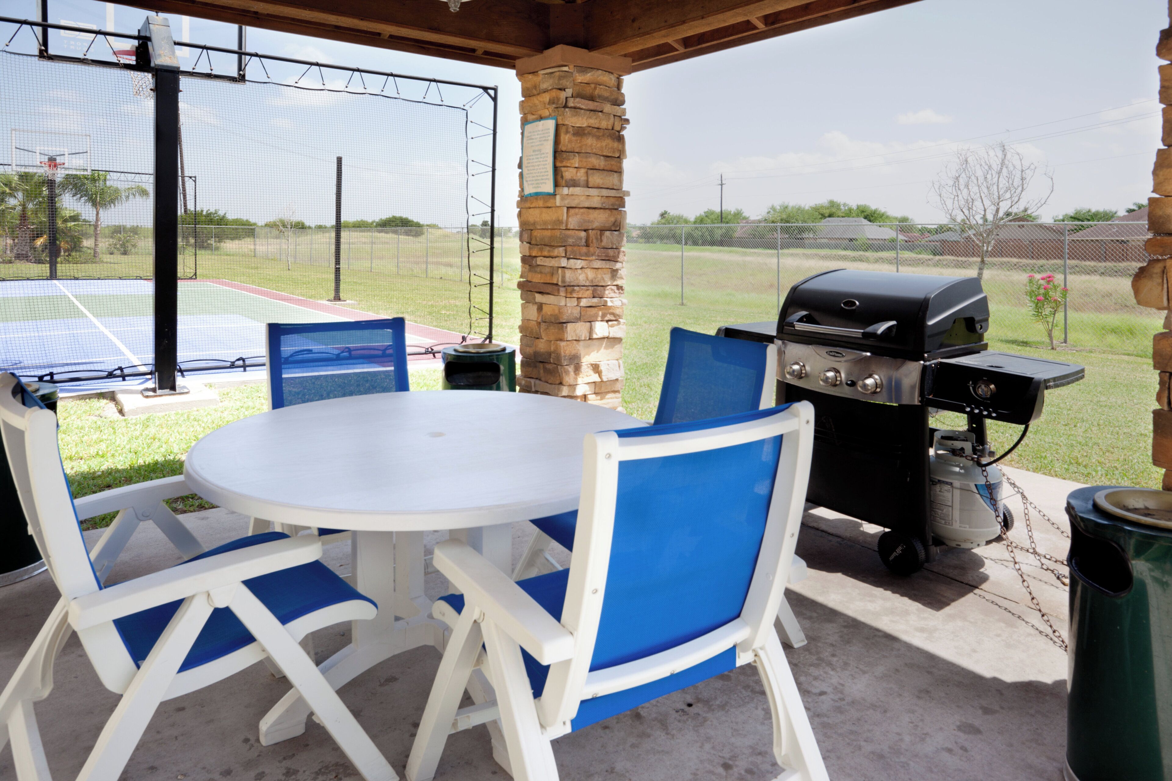 An outdoor patio with a round table and chairs, a barbecue grill, and a tennis court in the background.