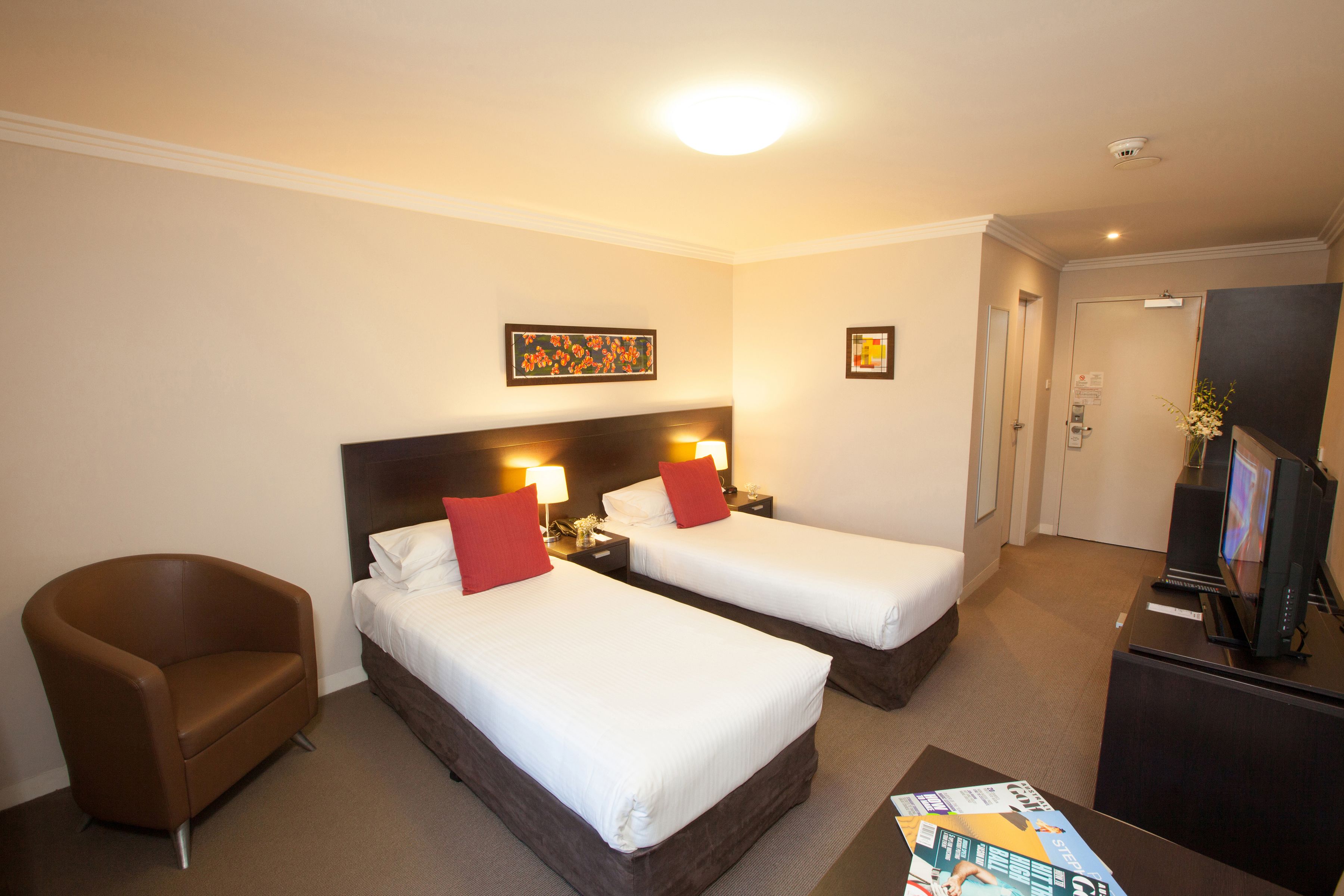 A hotel room with two single beds, a brown armchair, a desk with a television, and a magazine on the table.