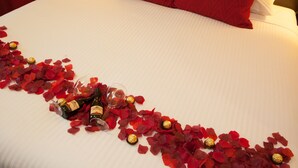 Een bed met rode rozenblaadjes en chocolade, een fles wijn en een glas op een wit laken.