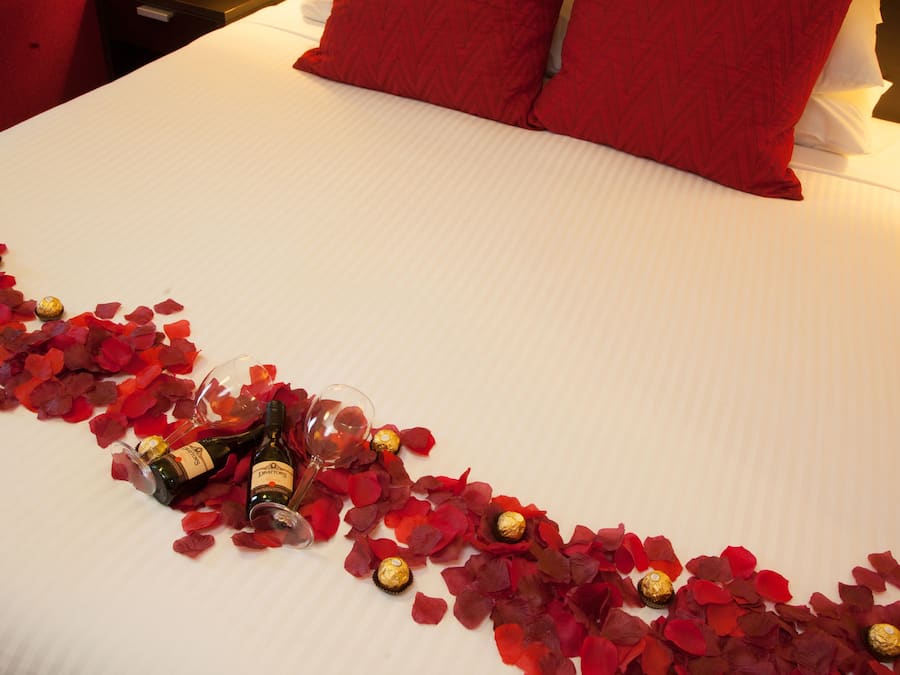 A bed with red rose petals and chocolates, a bottle of wine, and a glass on a white bedspread.