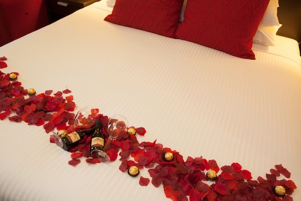 Una cama con pétalos de rosa roja y bombones, una botella de vino y una copa sobre un edredón blanco.