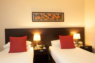 Habitación de hotel con dos camas, cada una con un almohadón rojo y ropa de cama blanca, una mesita de noche con una lámpara, un teléfono y una obra de arte enmarcada sobre las camas.