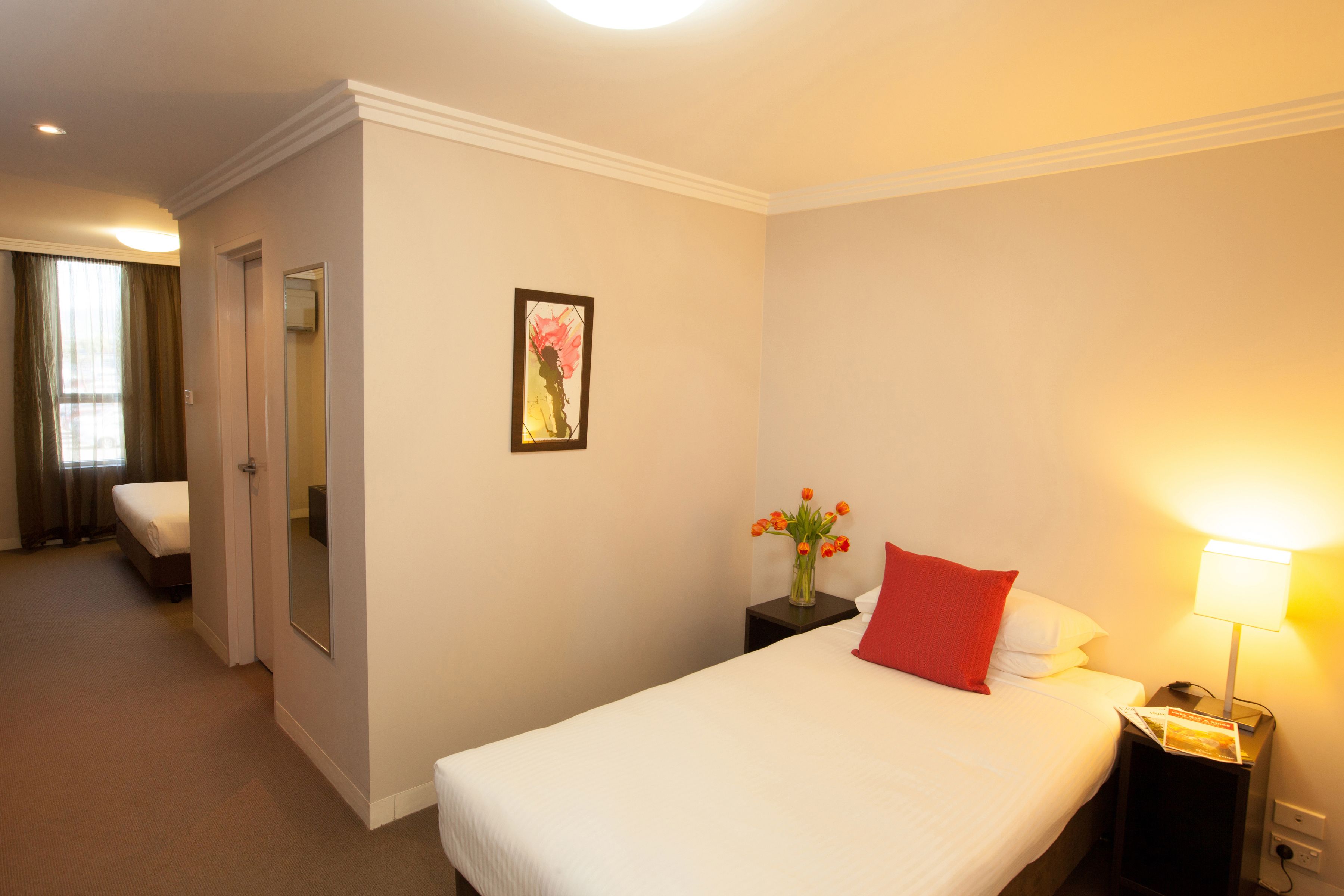 A hotel room with a bed, a nightstand with a lamp, a vase with flowers, a mirror, and a door leading to another room.